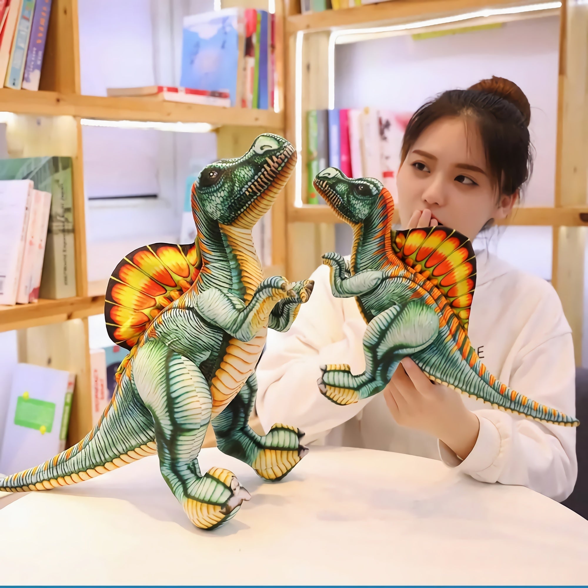 A person holds two colorful Spinosaurus plush toys while sitting at a table in a library setting.