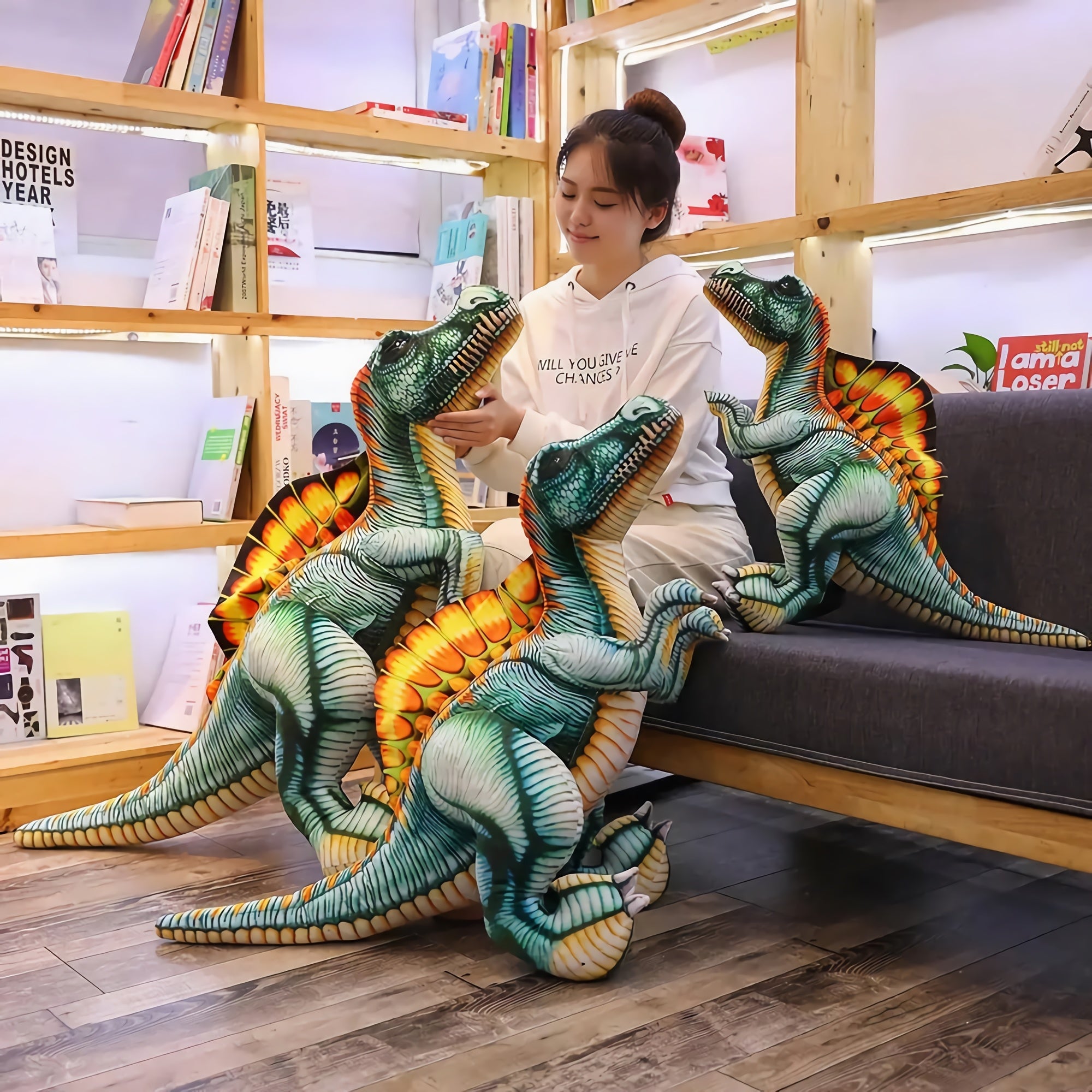 A person interacts with three colorful Spinosaurus plush pillows in a cozy room filled with books.