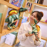 A person holds a small Spinosaurus plush toy while standing next to a larger one on a wooden shelf filled with books.