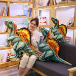 A woman sits on a couch holding a large Spinosaurus plush pillow, surrounded by smaller Spinosaurus plush toys.